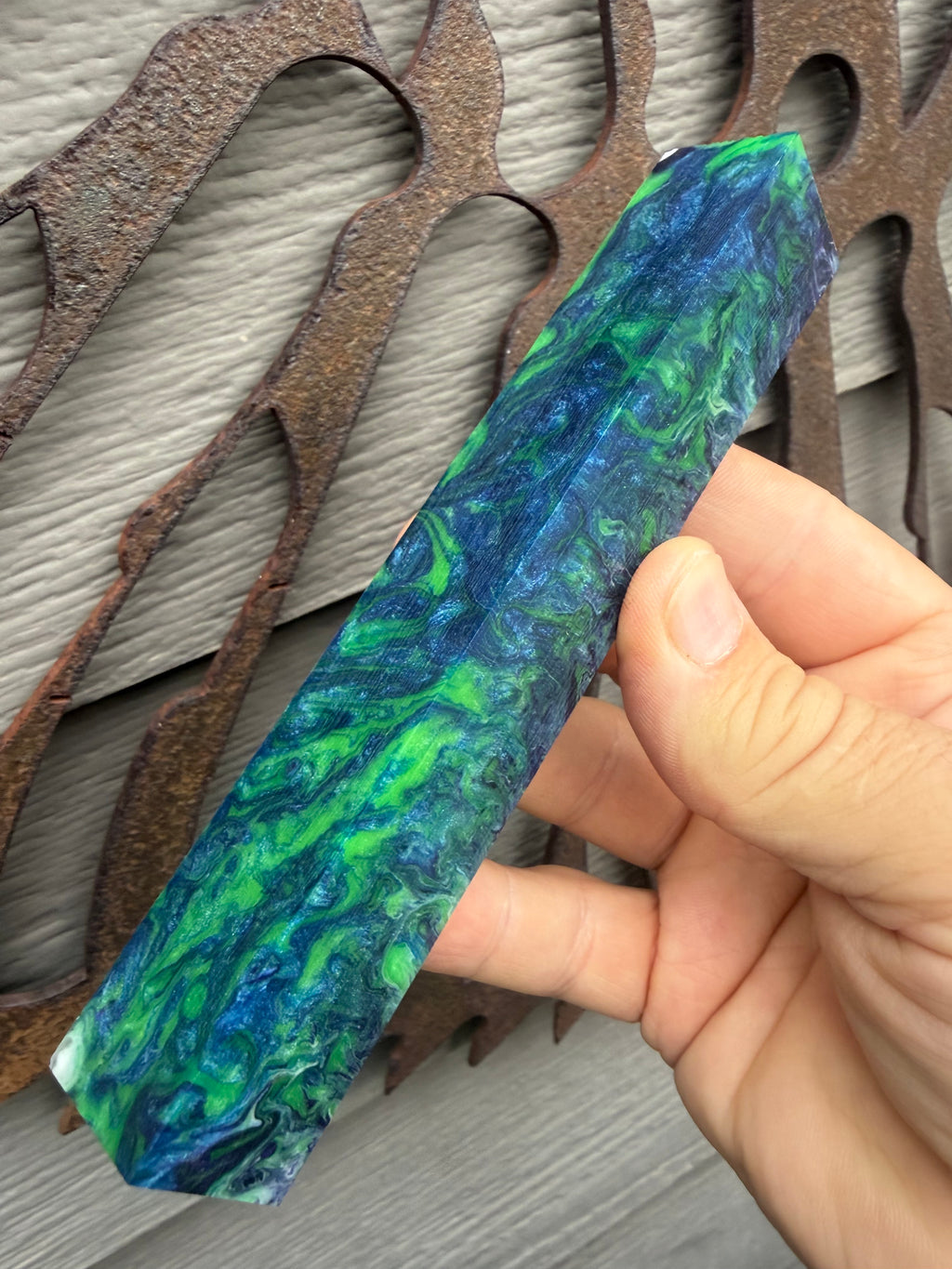 Resin pen and bead blanks for wood andnl lathe turners A hand holding a long, rectangular marbled resin or glass rod with swirls of blue, green, and teal, set against a background of rustic metal cutouts and gray siding.
