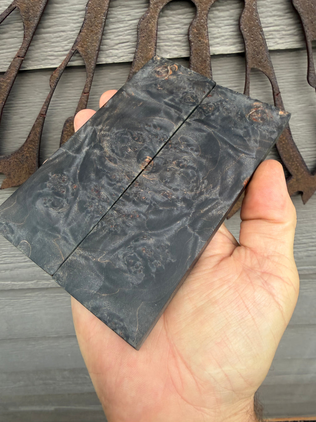 A hand holding a dark, polished piece of burl wood with dramatic swirling grain patterns, set against a background of rustic metal cutouts on a gray wood siding wall. Stabilized maple Burl knife scales 